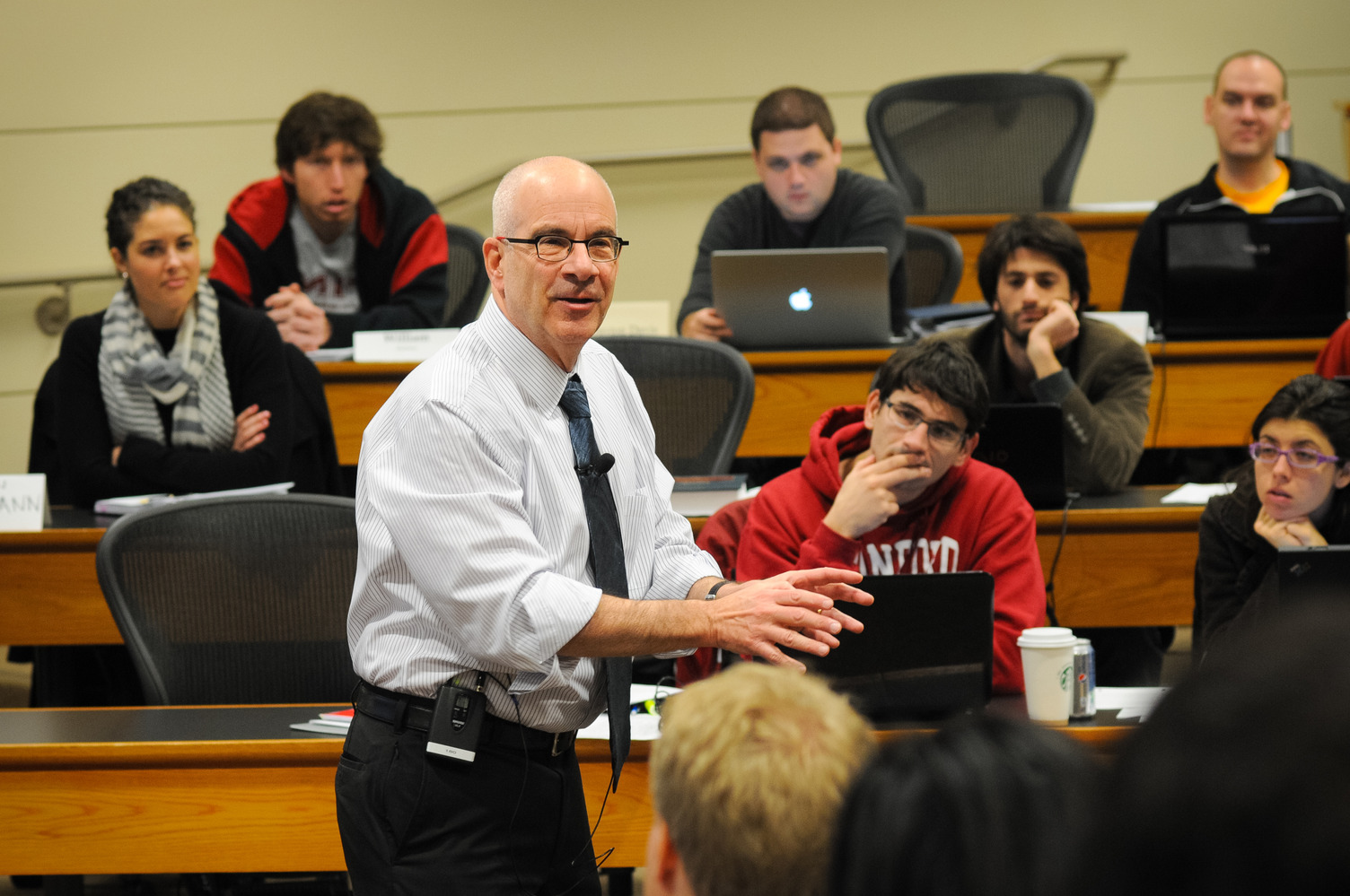 Joseph Grundfest gestures while speaking to students seated at desks with laptops in a classroom setting, engaging the audience during a lecture.