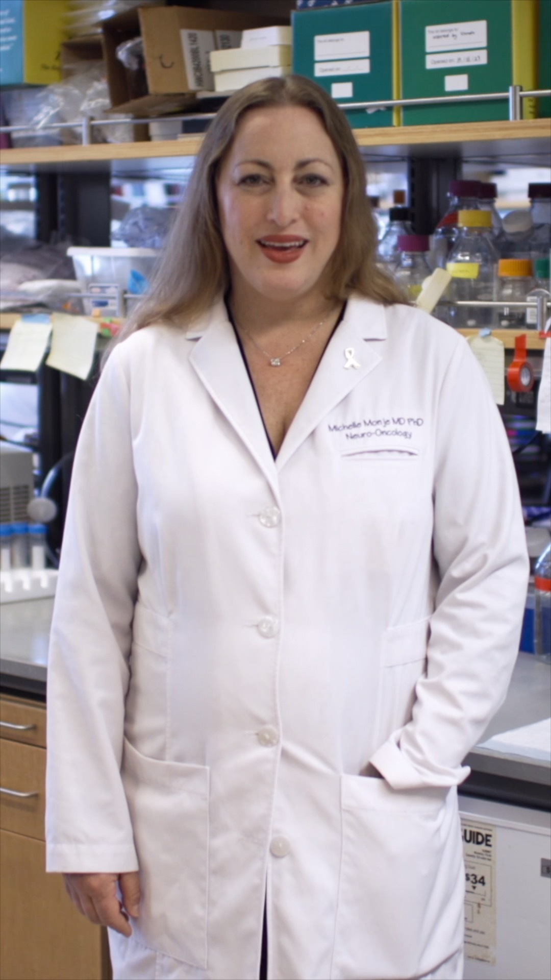 Michelle Monje in a lab coat stands in a laboratory filled with equipment and materials related to scientific research.