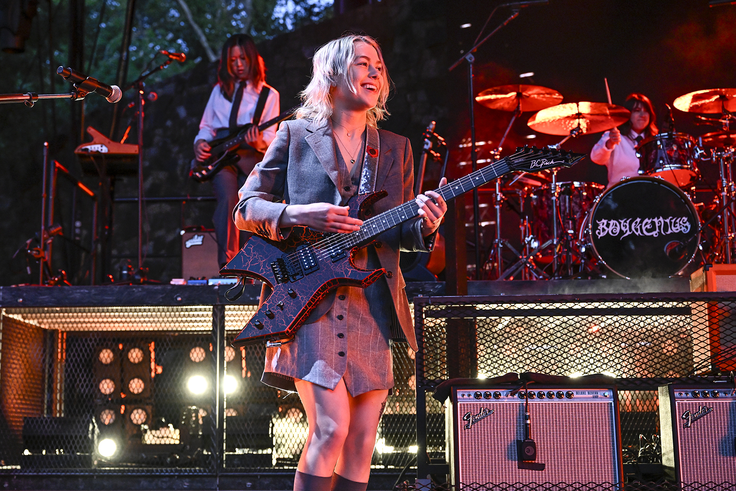 Phoebe Bridgers performing on stage in a stylish outfit, with a drummer and band members visible in the background.