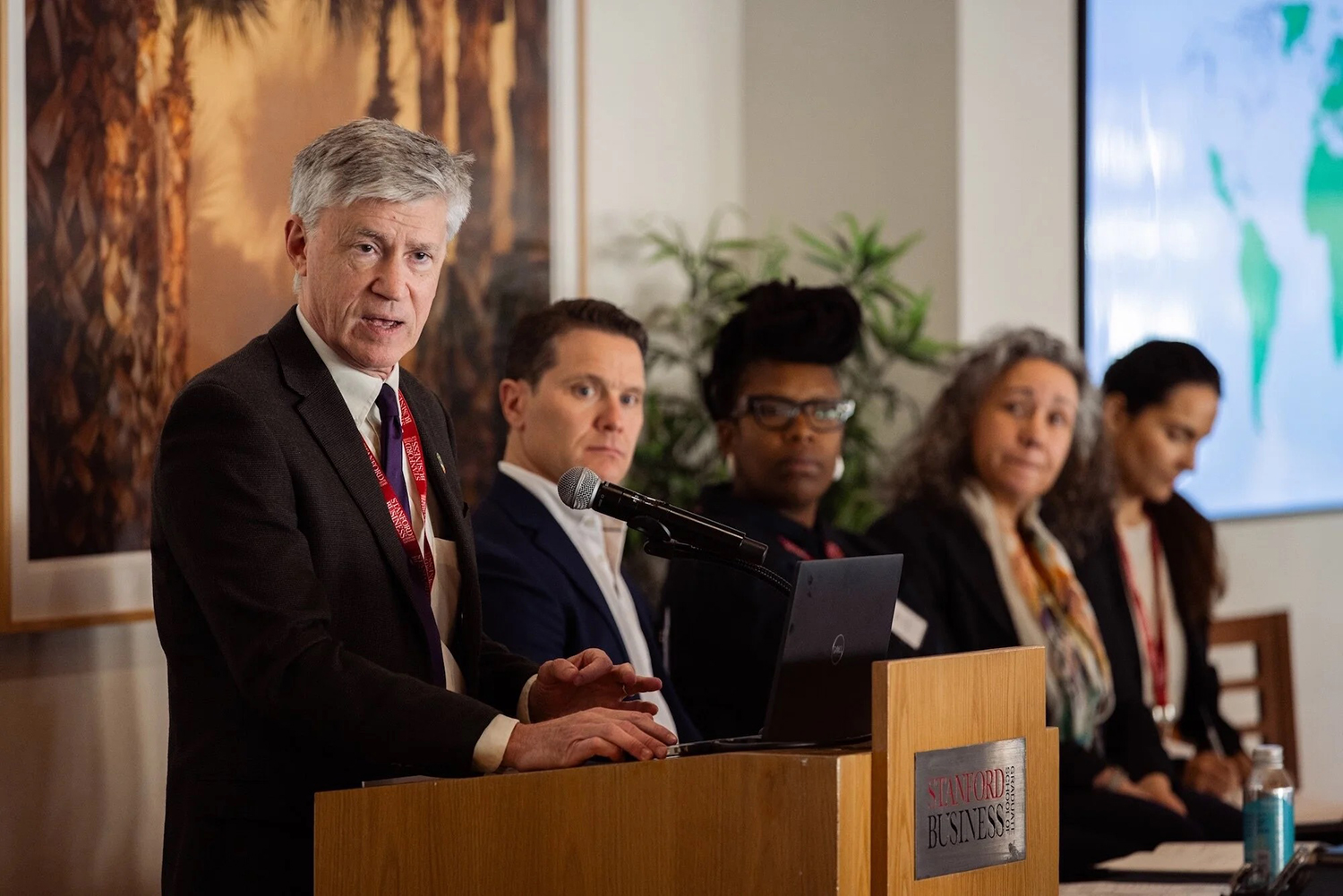 John Knox addresses an audience at a podium in front of a world map, with panelists visible in the background.