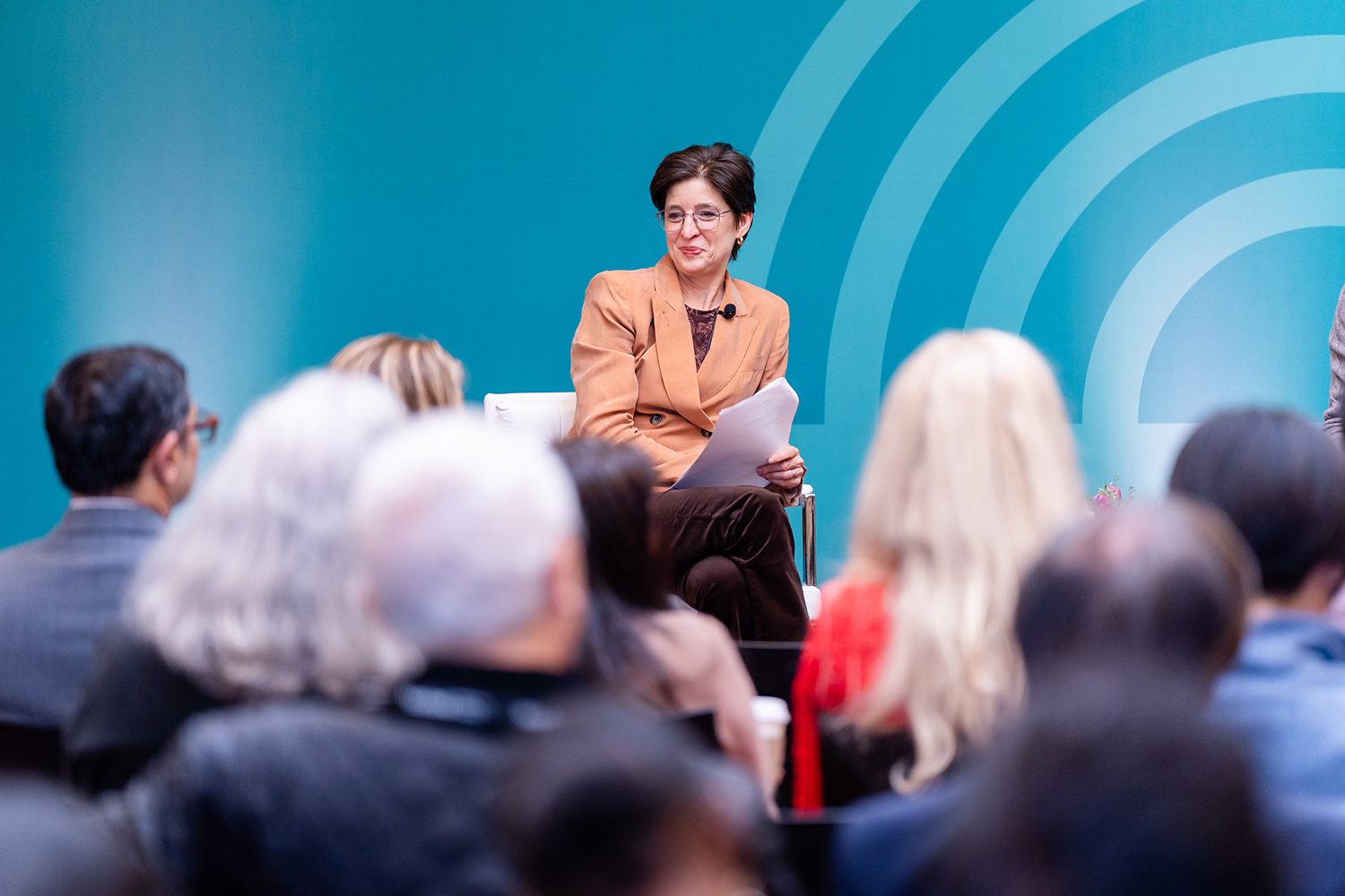 A speaker holds notes while addressing an audience at an event, set against a blue background.