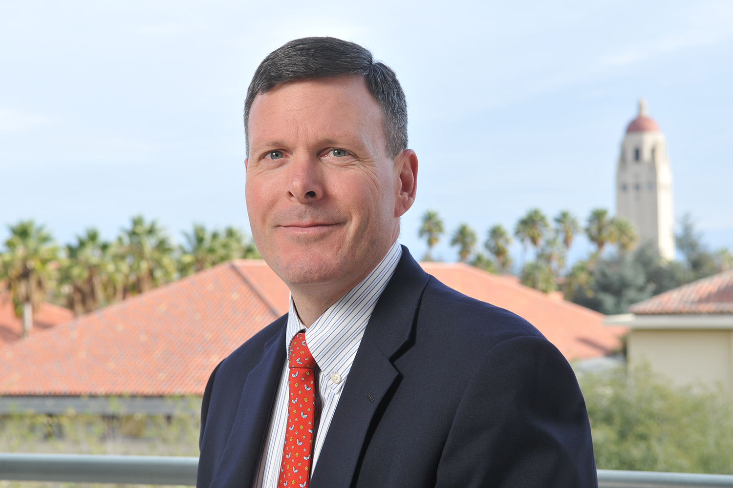 Calvert in suit and red tie standing outdoors with palm trees and a Hoover Tower in the background.