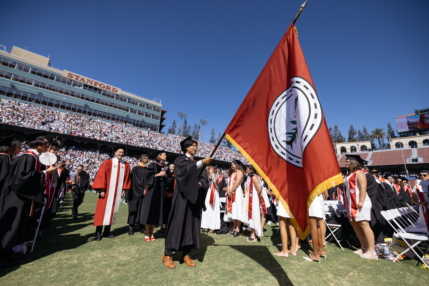 Graduate carries a large red flag with the presidential emblem during outdoor stadium commencement ceremony.
