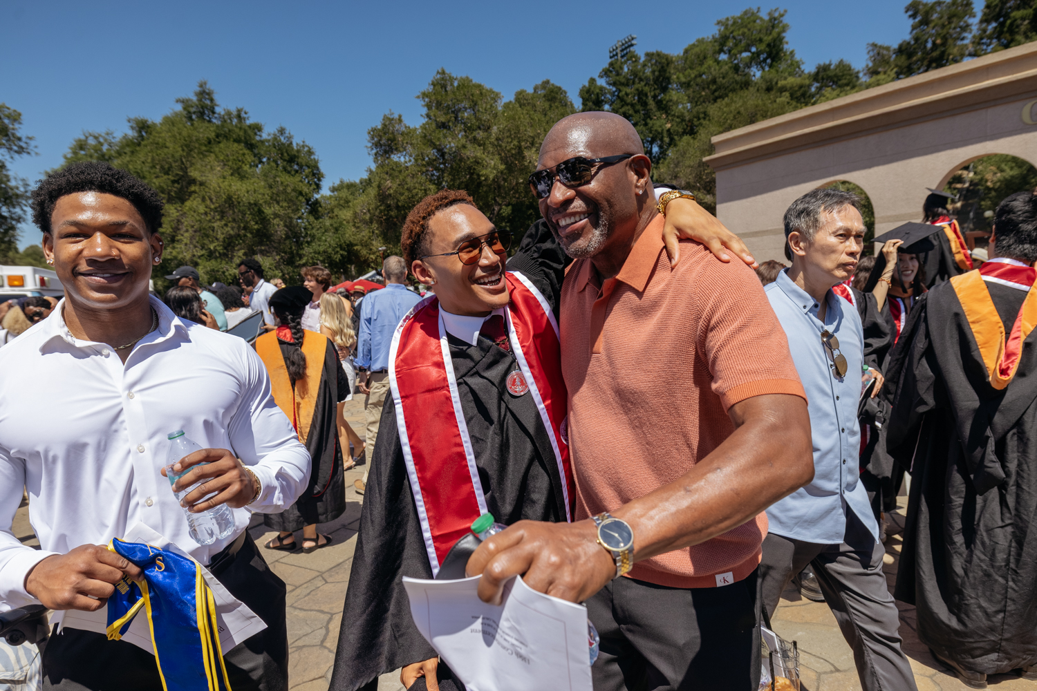 Graduate in cap and gown embraces family member after outdoor commencement ceremony.
