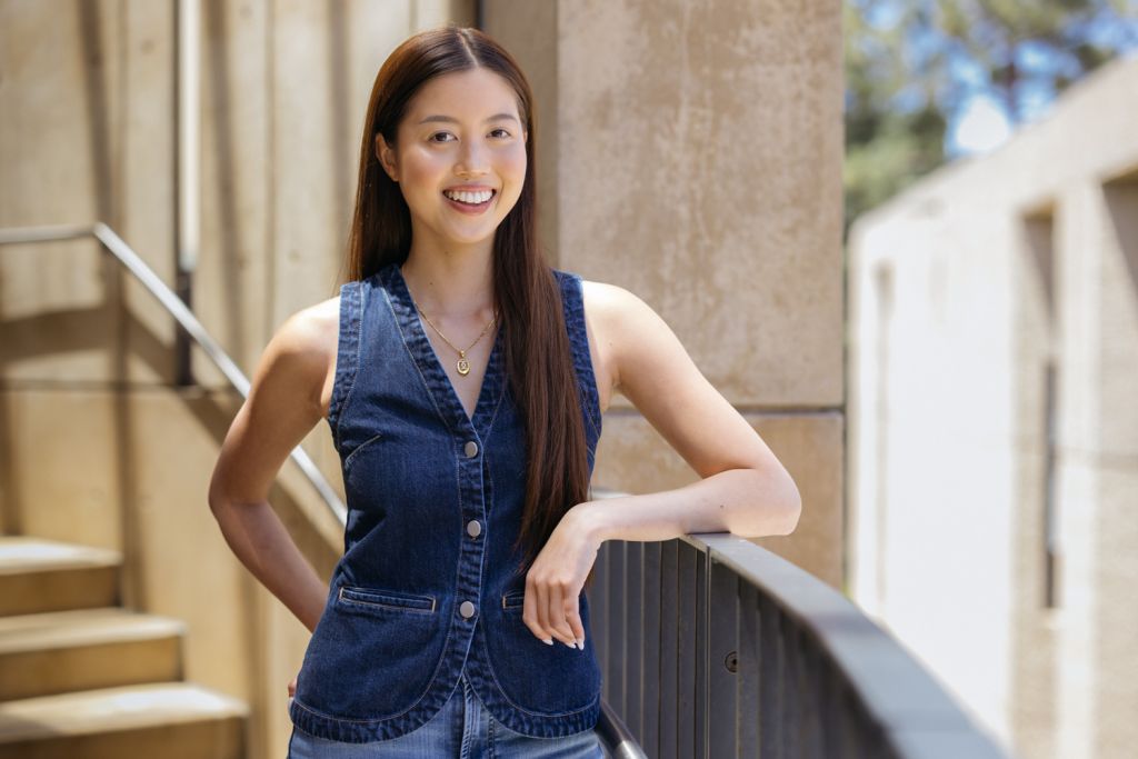 Chung with long hair and smiling in a denim vest stands near stone columns.