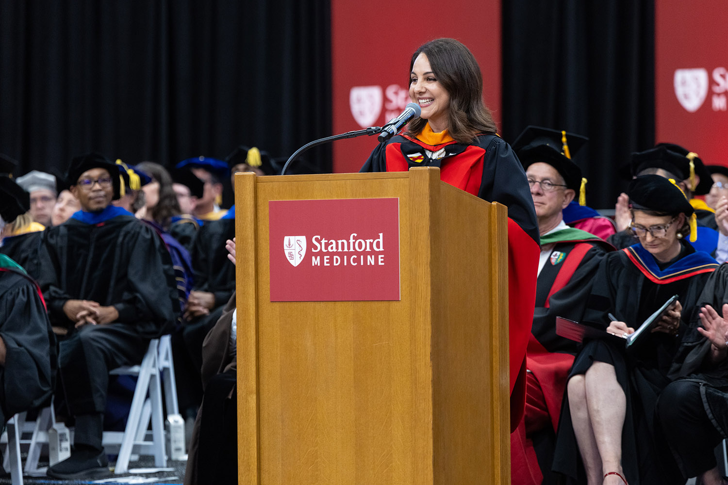 Keynote speaker Lily Sarafan in academic regalia addresses graduates from podium at Stanford Medicine commencement ceremony.