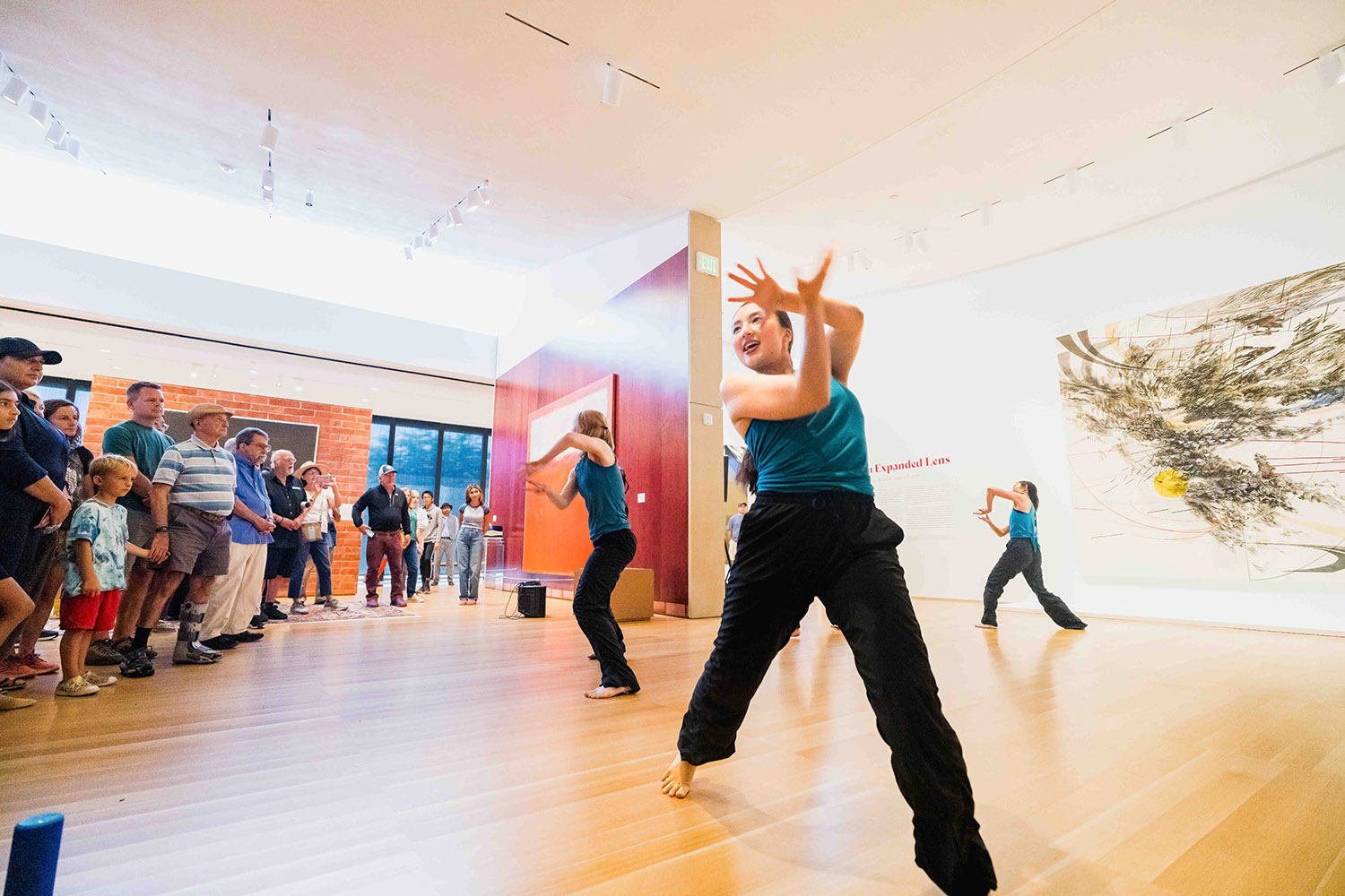 Dancers perform in art gallery as an audience watches, with colorful artwork displayed on the walls behind them.