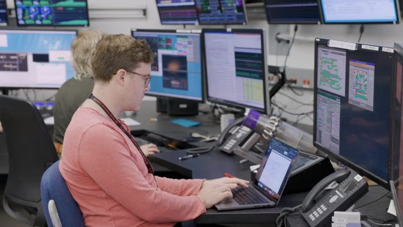 A man works at a control desk with multiple computer monitors displaying data and graphs, focused on their laptop.