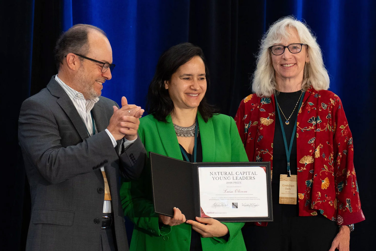 A woman in a green jacket holds a certificate, smiling with two people beside her during the Natural Capital Young Leaders award ceremony.