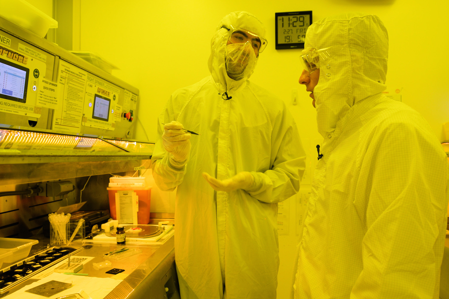 Open video Two people in protective gear discuss procedures near specialized lab equipment, with various tools and documents visible on the table. in a modal