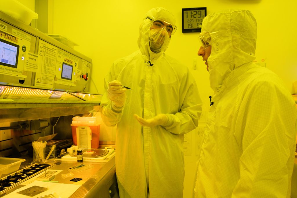 Open video Two people in protective gear discuss procedures near specialized lab equipment, with various tools and documents visible on the table. in a modal