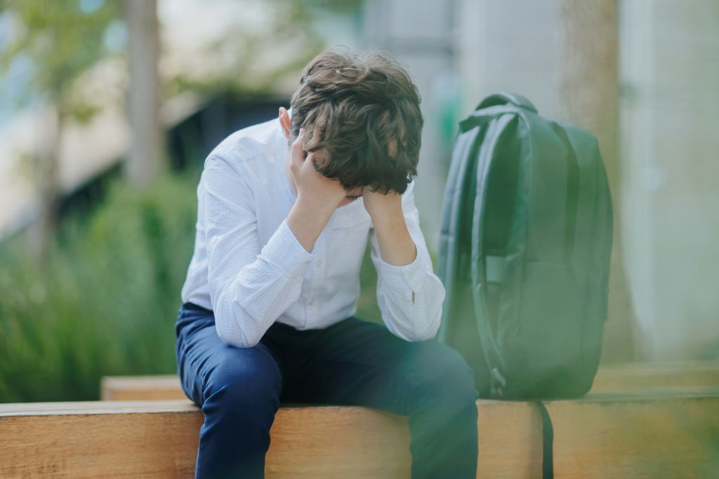 A young boy sits on a bench with his head in his hands, looking distressed, with a backpack beside him.