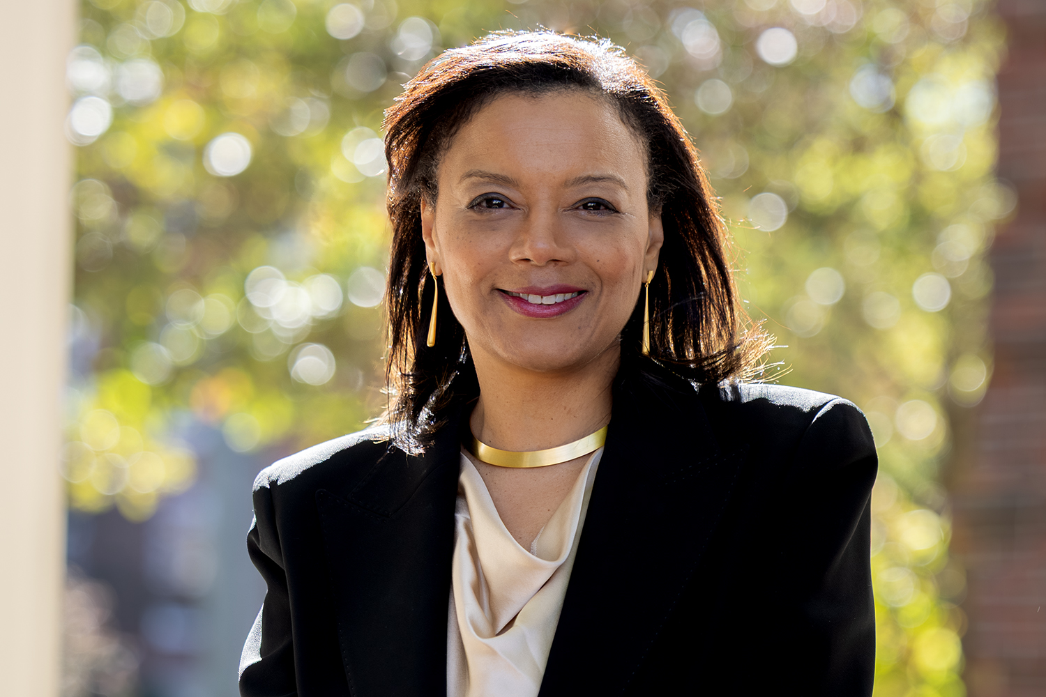 Woman in a black blazer and gold necklace smiling outdoors with blurred trees in the background.