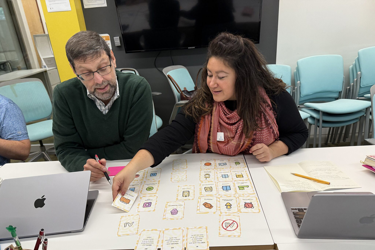 Two people collaborate at a table, examining cards and notes while using laptops in a classroom setting.