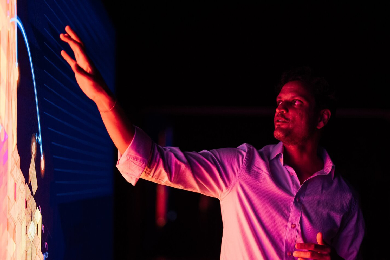 A man reaches toward a glowing interactive screen showing colorful digital patterns in a dark room.