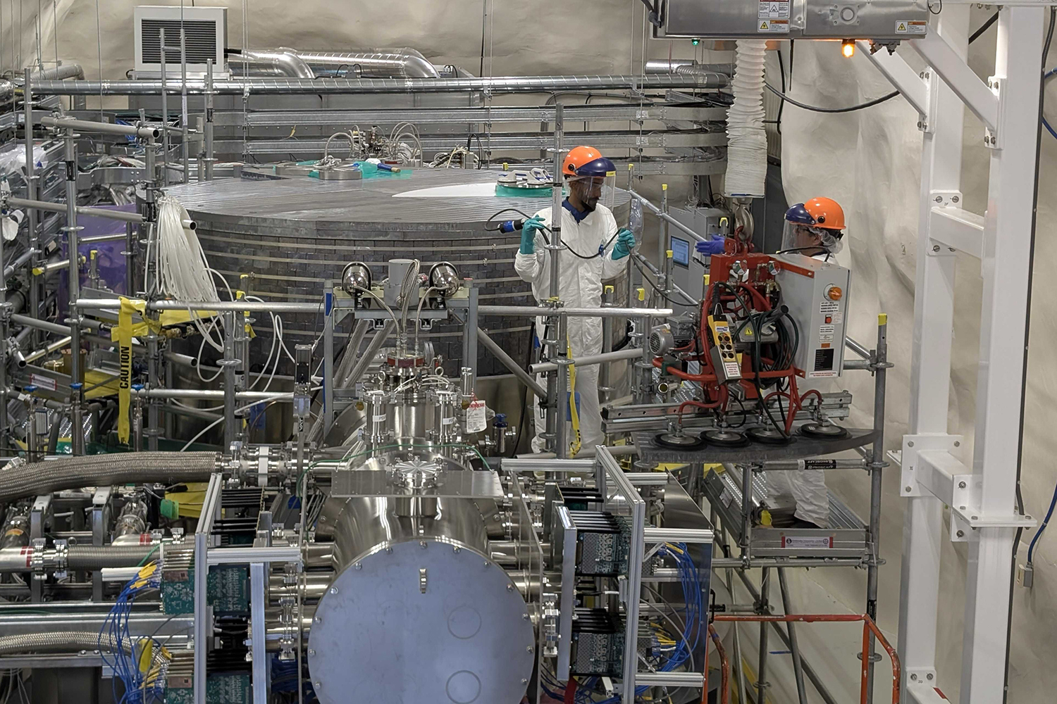 Two workers in white coveralls and orange hard hats work on scaffolding beside large stainless steel tanks and piping.