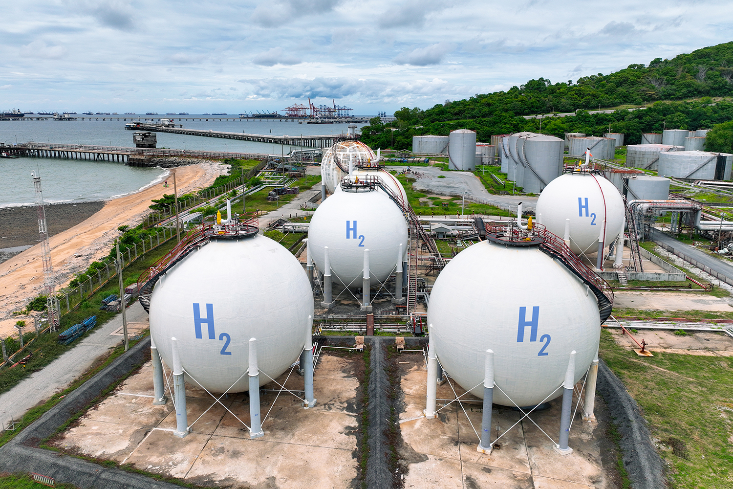 Large spherical tanks store hydrogen gas at a coastal industrial plant, with a shipping port visible in the background.