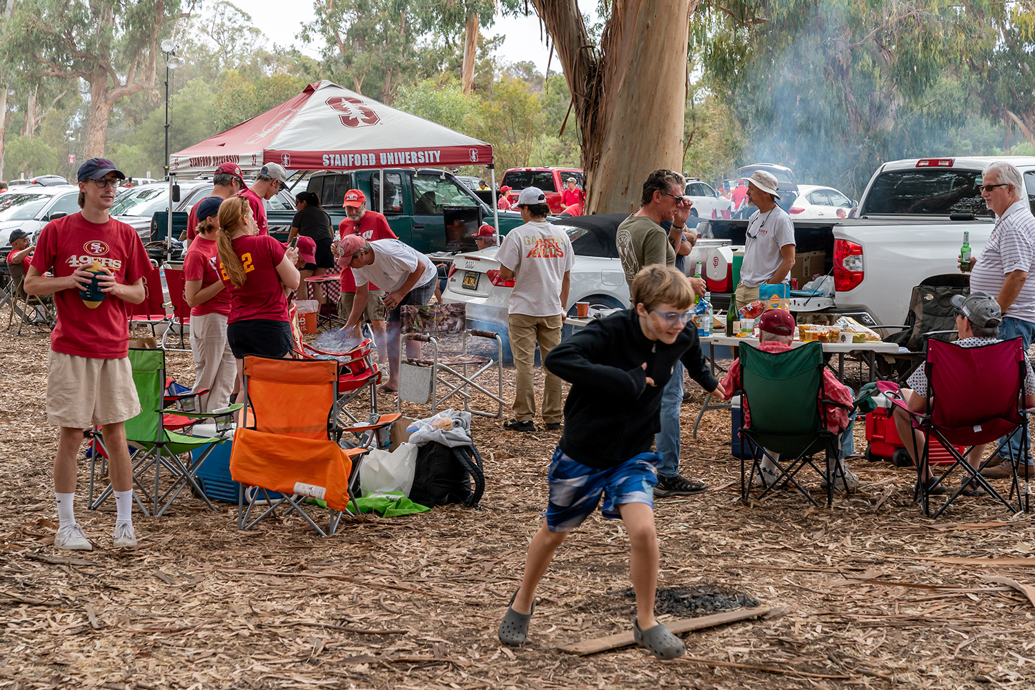 Families tailgate under eucalyptus trees, grilling food and setting up camp chairs before a football game.