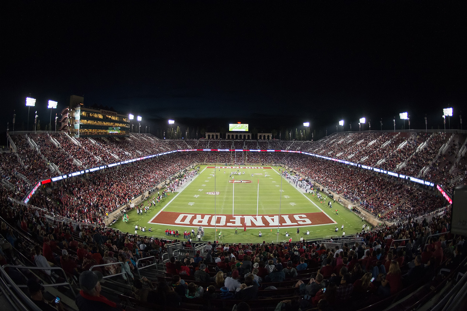An aerial view shows Stanford Stadium packed with fans during a night football game.