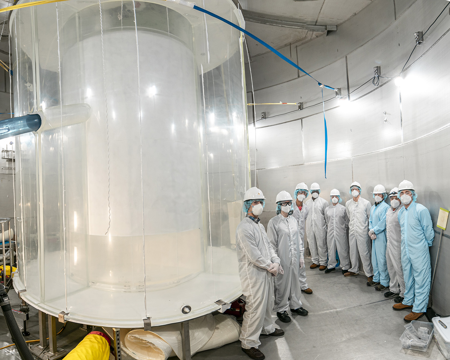 A team in clean suits and hard hats stands beside a tall transparent cylindrical detector tank.