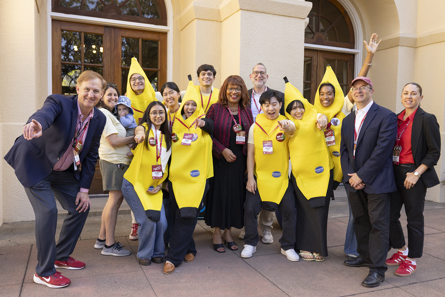 University staff and students in banana costumes pose together in front of a campus building entrance.