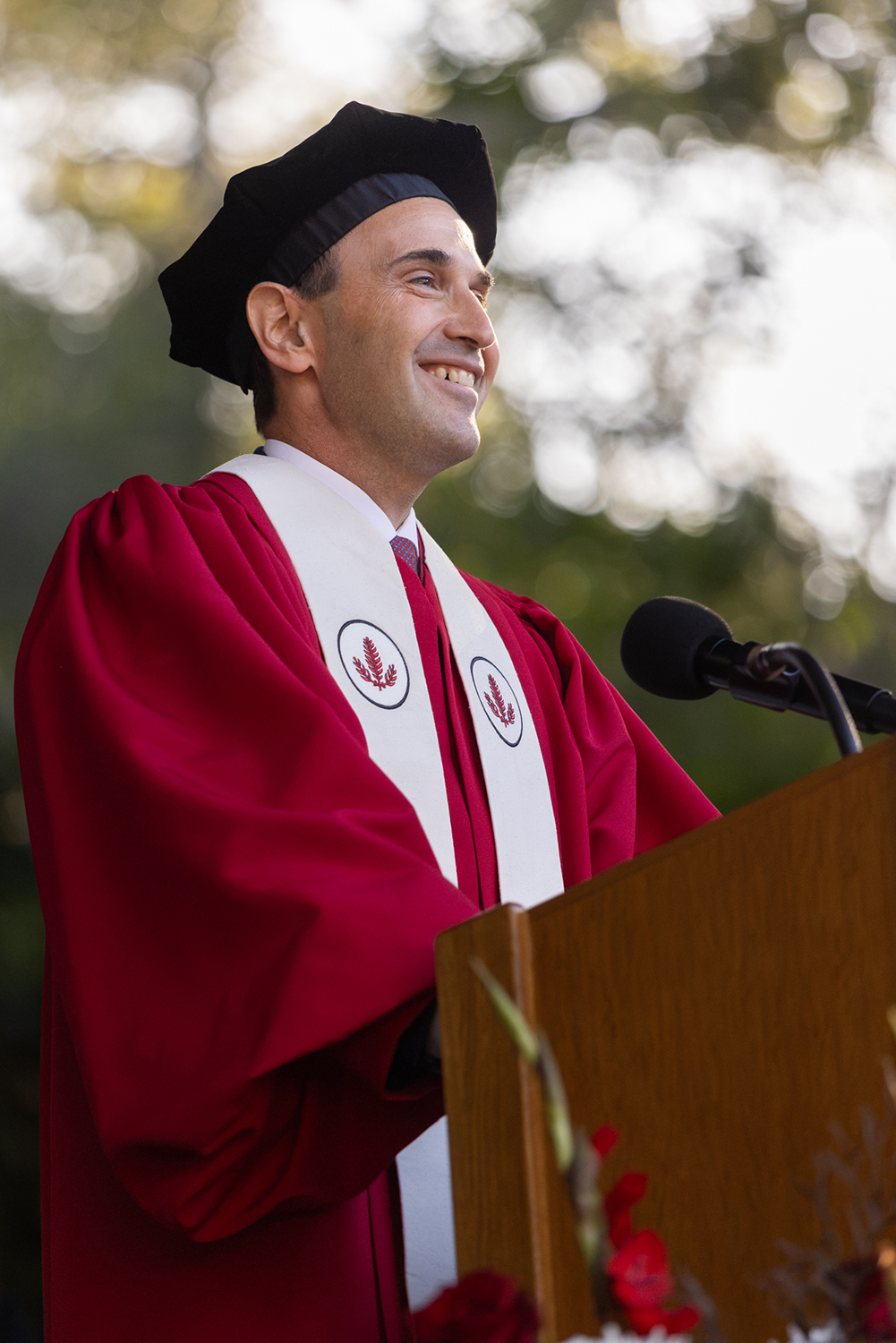 President Jonathan Levin wearing academic regalia smiles while speaking at a podium outdoors.