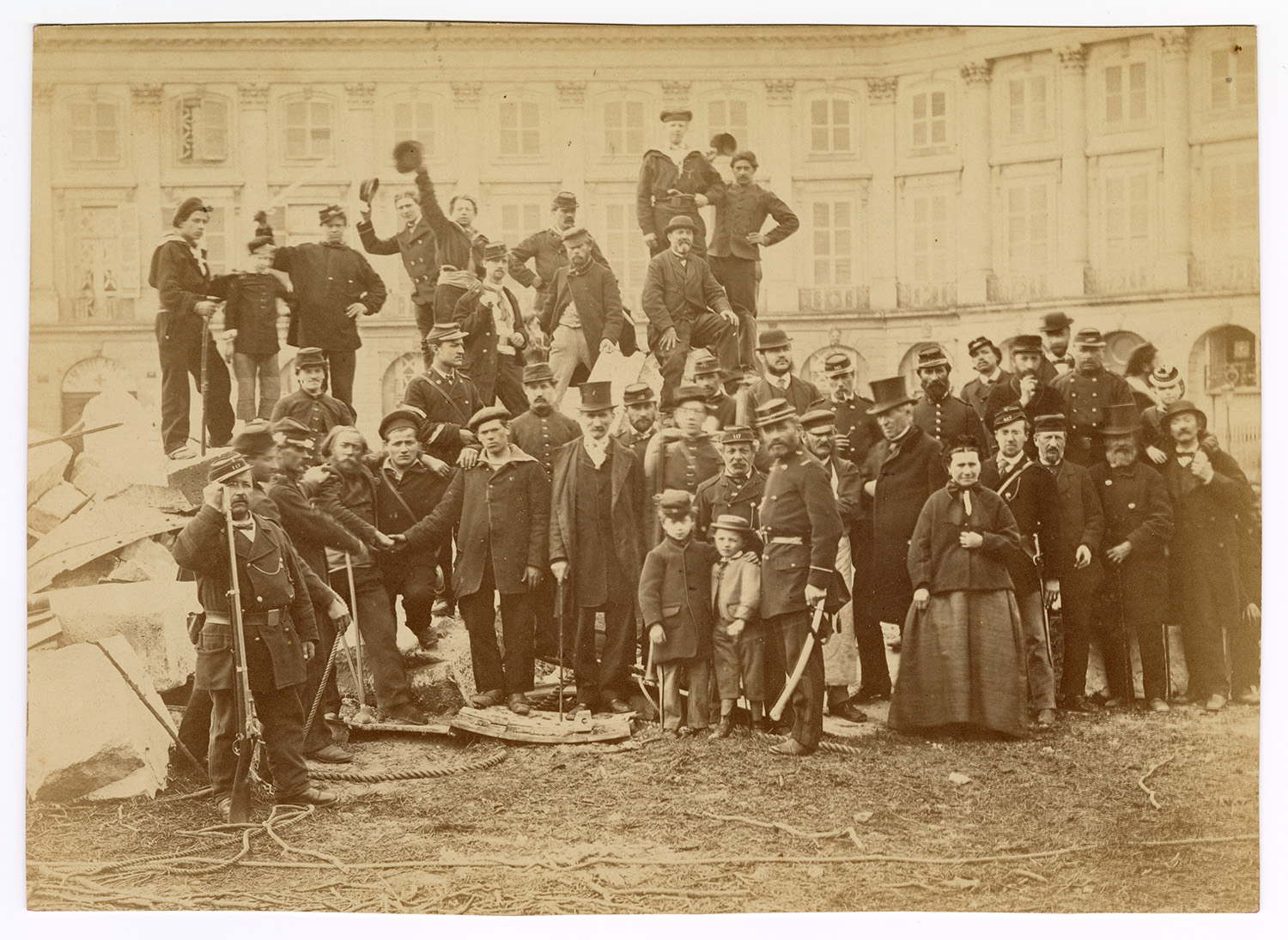 An archival photo captures a large group posed atop rubble in a Paris square, some raising their hats for the photograph.