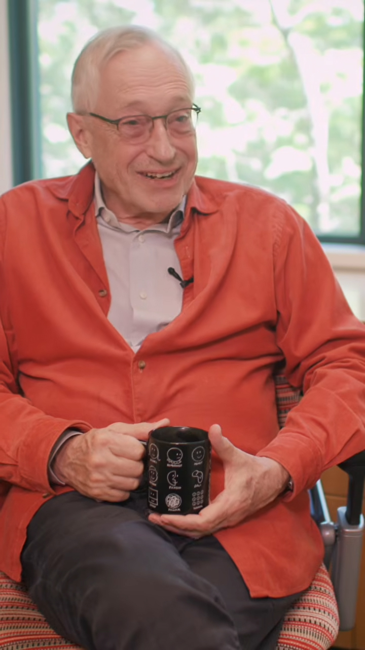 Paul Milgrom holds a black mug while seated by a window, wearing a small clip-on microphone for an interview.