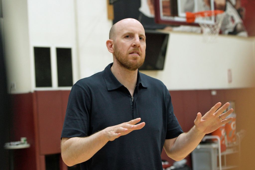 Dan Grunfeld gestures with both hands while speaking in a gym with a basketball hoop behind him.