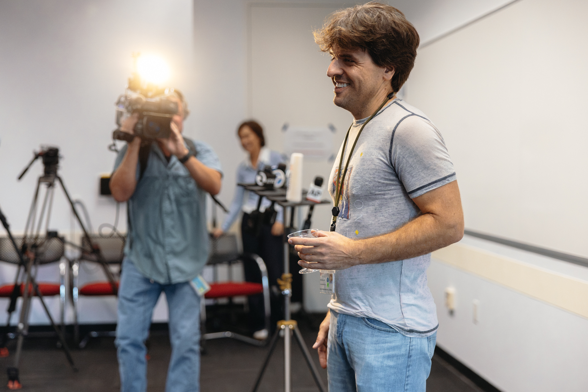 Karl Deisseroth holding a plastic champagne coupe stands near news microphones as a TV camera operator films.