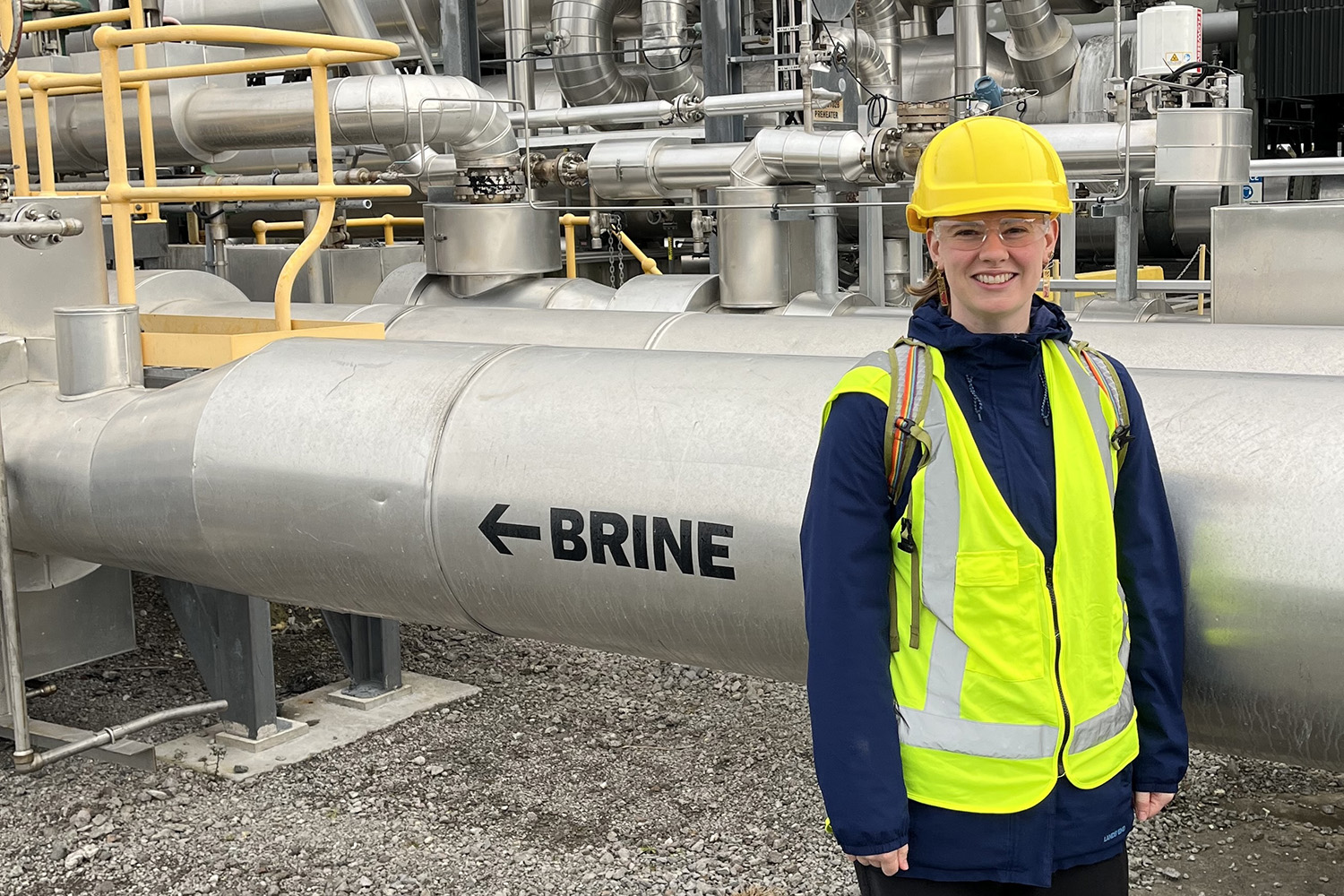 Laura Blackstone in a hard hat, safety glasses, and reflective vest stands beside a large pipe labeled 