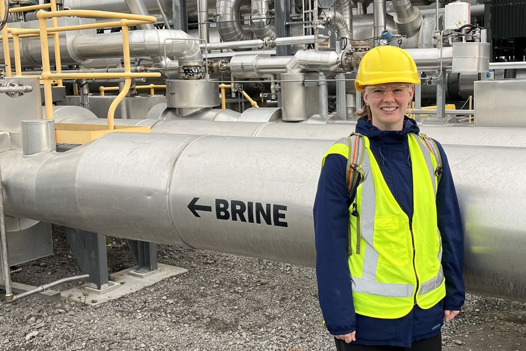 Laura Blackstone in a hard hat, safety glasses, and reflective vest stands beside a large pipe labeled 