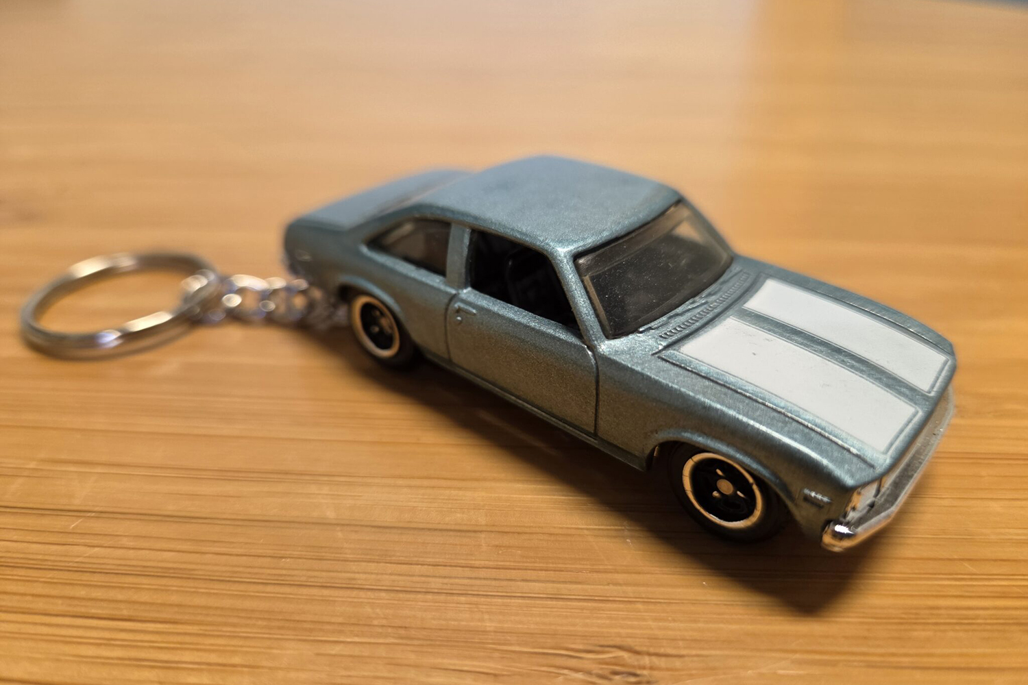 A gray-blue toy car with white hood stripes, attached to a metal key ring, sits on a wooden surface.