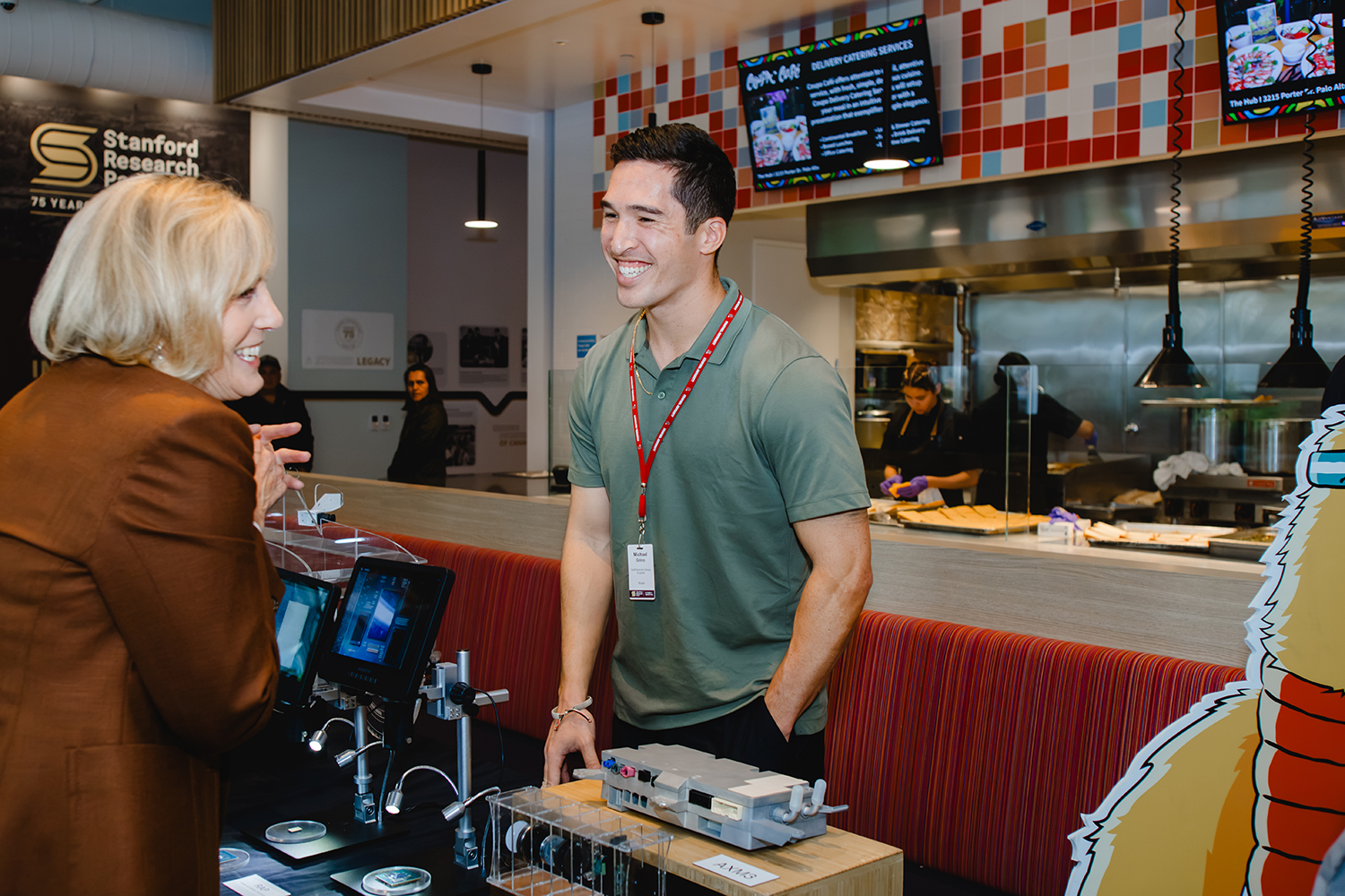 A smiling exhibitor in a green polo talks with a visitor at a table displaying electronic hardware and microscopes.