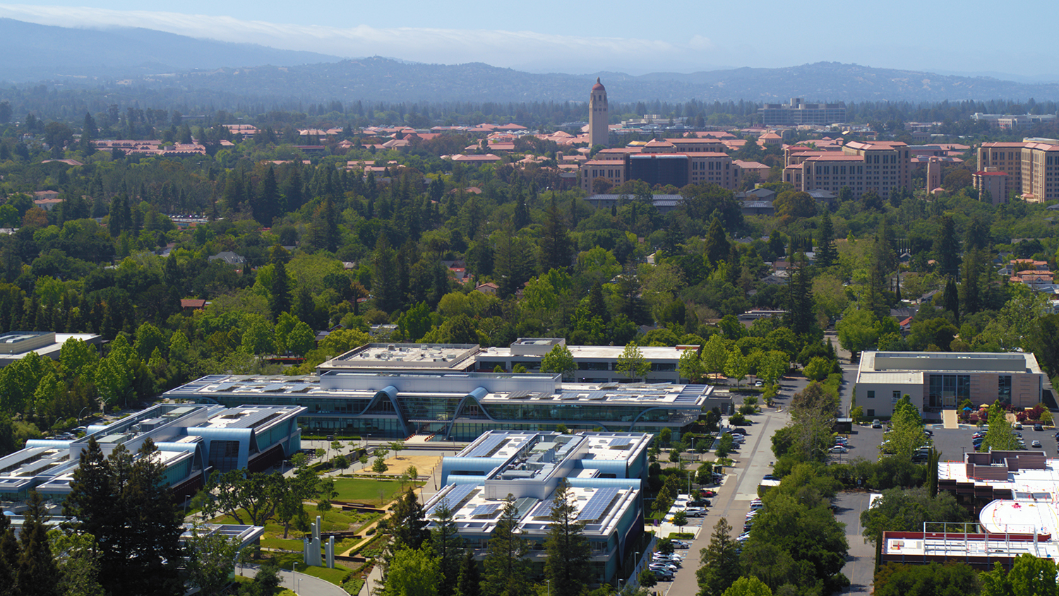 Aerial view of the modern, solar-paneled research park buildings, with Hoover Tower and the Stanford campus in the distance.