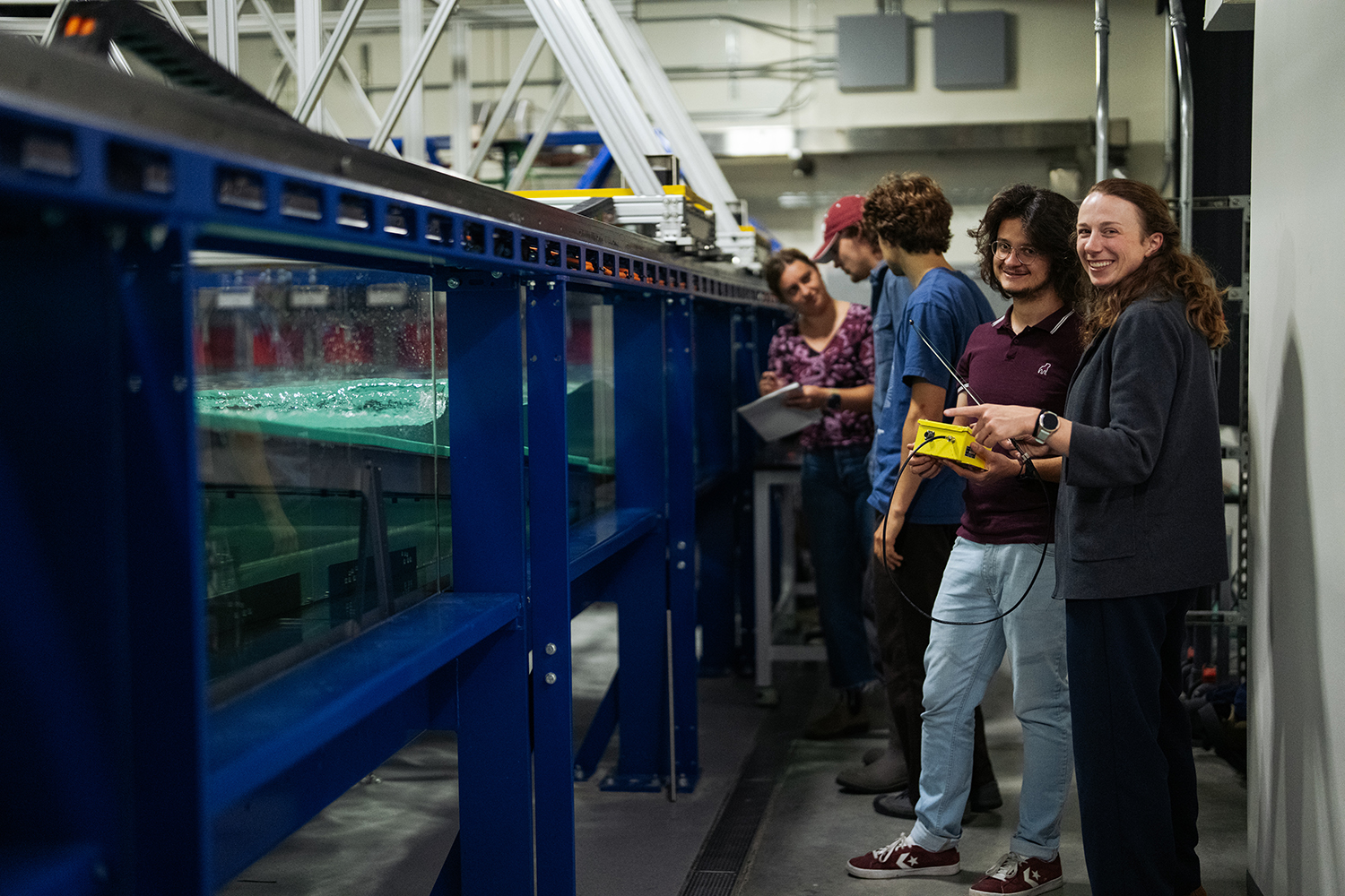 Lab members gather beside a long wave-generating flume tank, with two holding equipment and smiling toward the camera.