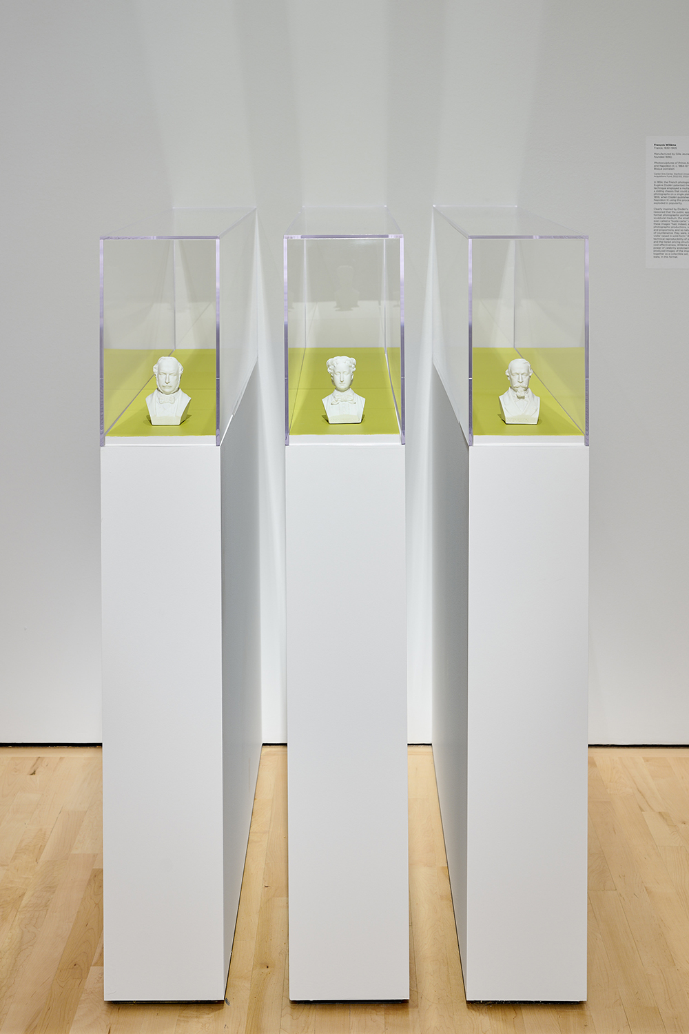 Three small white bust sculptures stand in separate glass display cases lit against a pale yellow background.