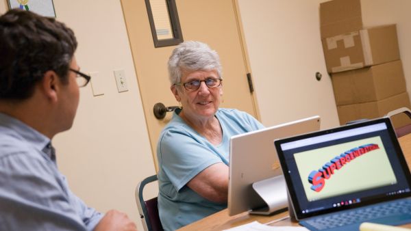 Young man talking with an older woman who sits in front of a computer screen. Logo for the project on another screen.