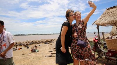 two women taking a selfie in front of thatched roof huts and the Mekong river