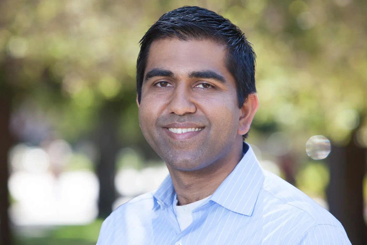 Neil Malhotra wearing a light blue shirt stands outdoors with a blurred background of trees.