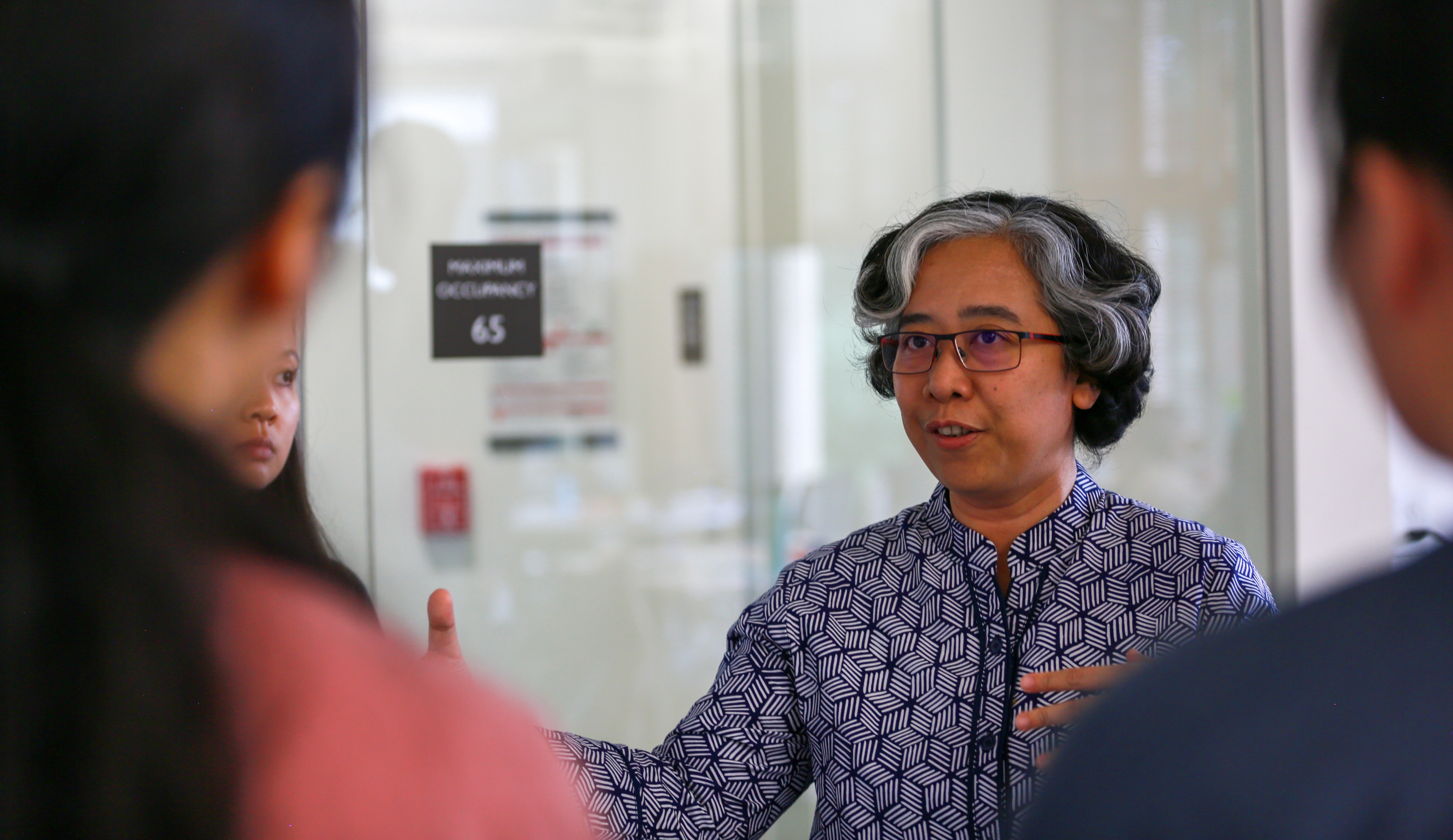 Dian Rositawati talking to a group of people. She is wearing a patterned shirt and glasses, and gesturing with her hands.
