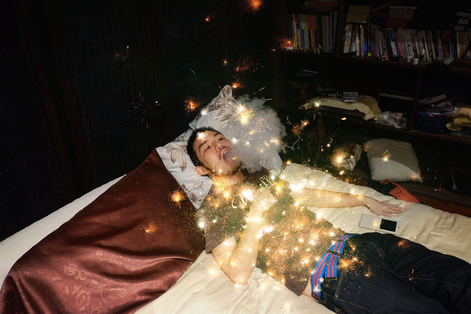 An edited photo work of a young person lying on a mattress in a dark room; the young person exhales a cloud of smoke. A bright scene of the cosmos is imposed over the person's body.