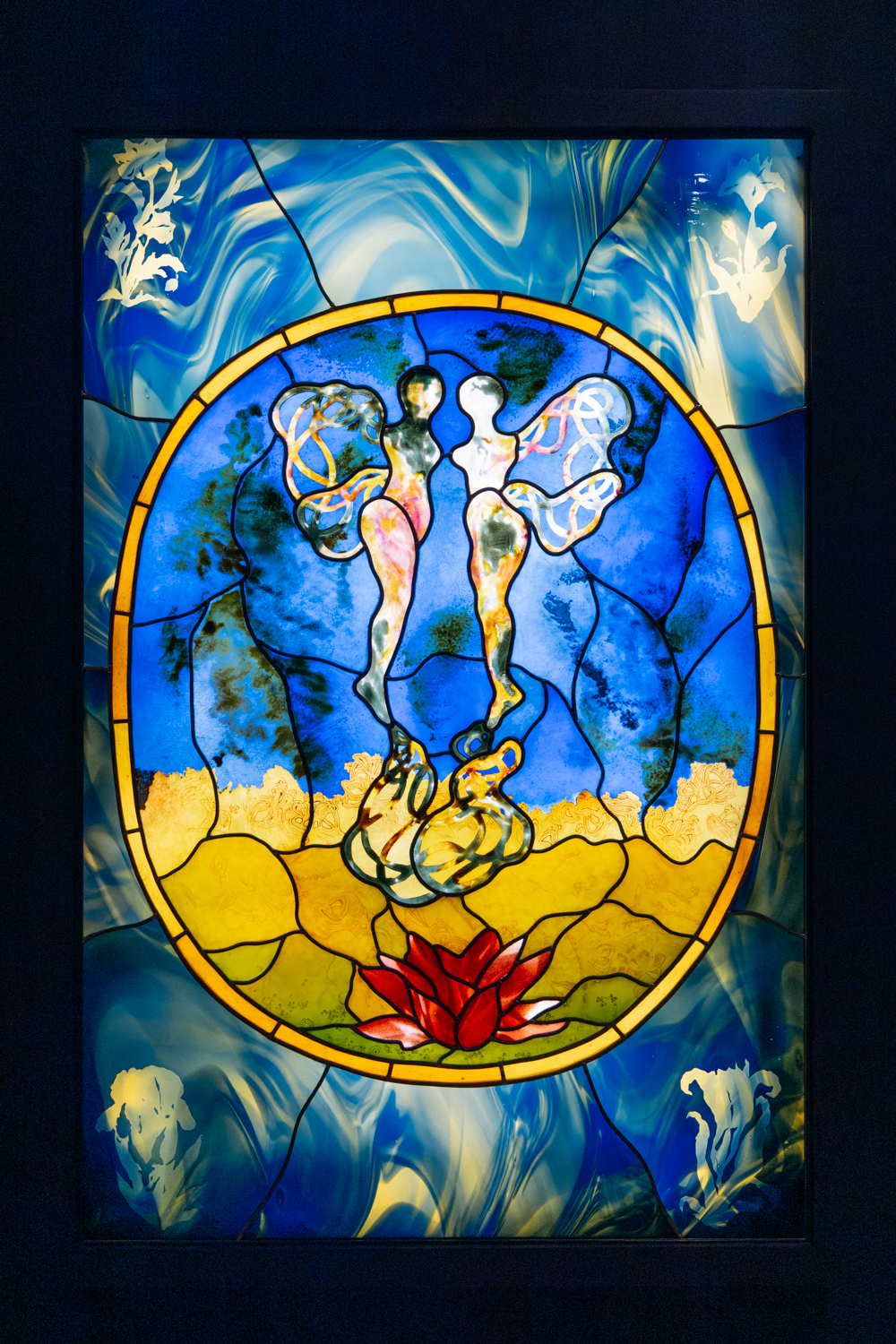 A glass artwork features vibrant colors and intricate designs, including figures in a dynamic pose against a blue and gold backdrop framed by an oval border. A red lotus flower is prominently displayed at the bottom.