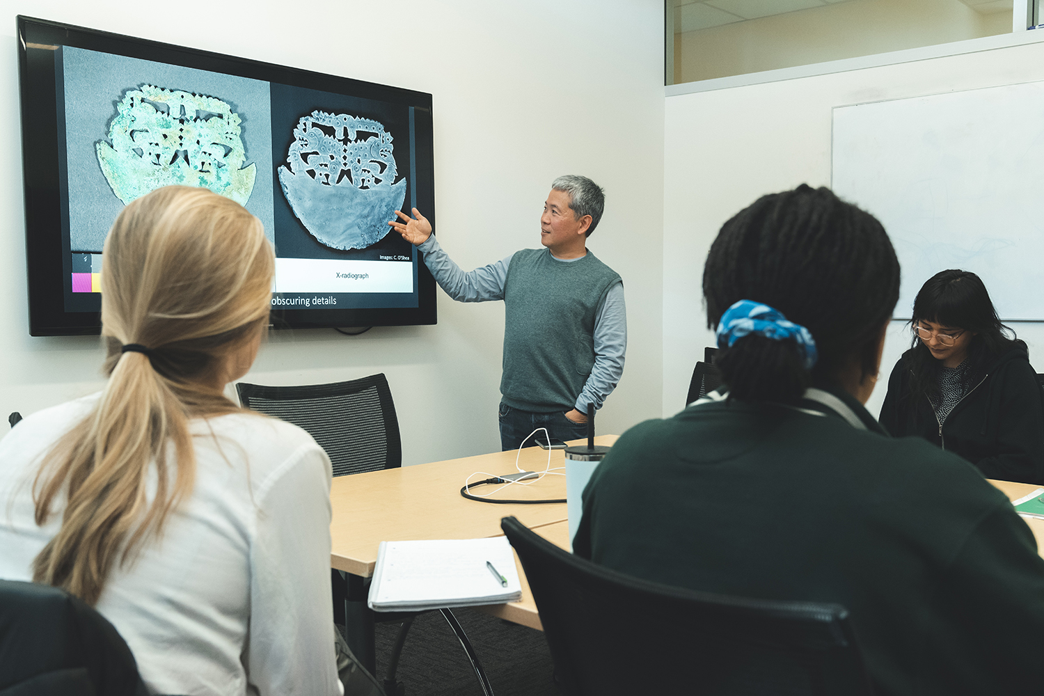 In a classroom, a presenter gestures toward two images on a screen while an attentive audience listens.