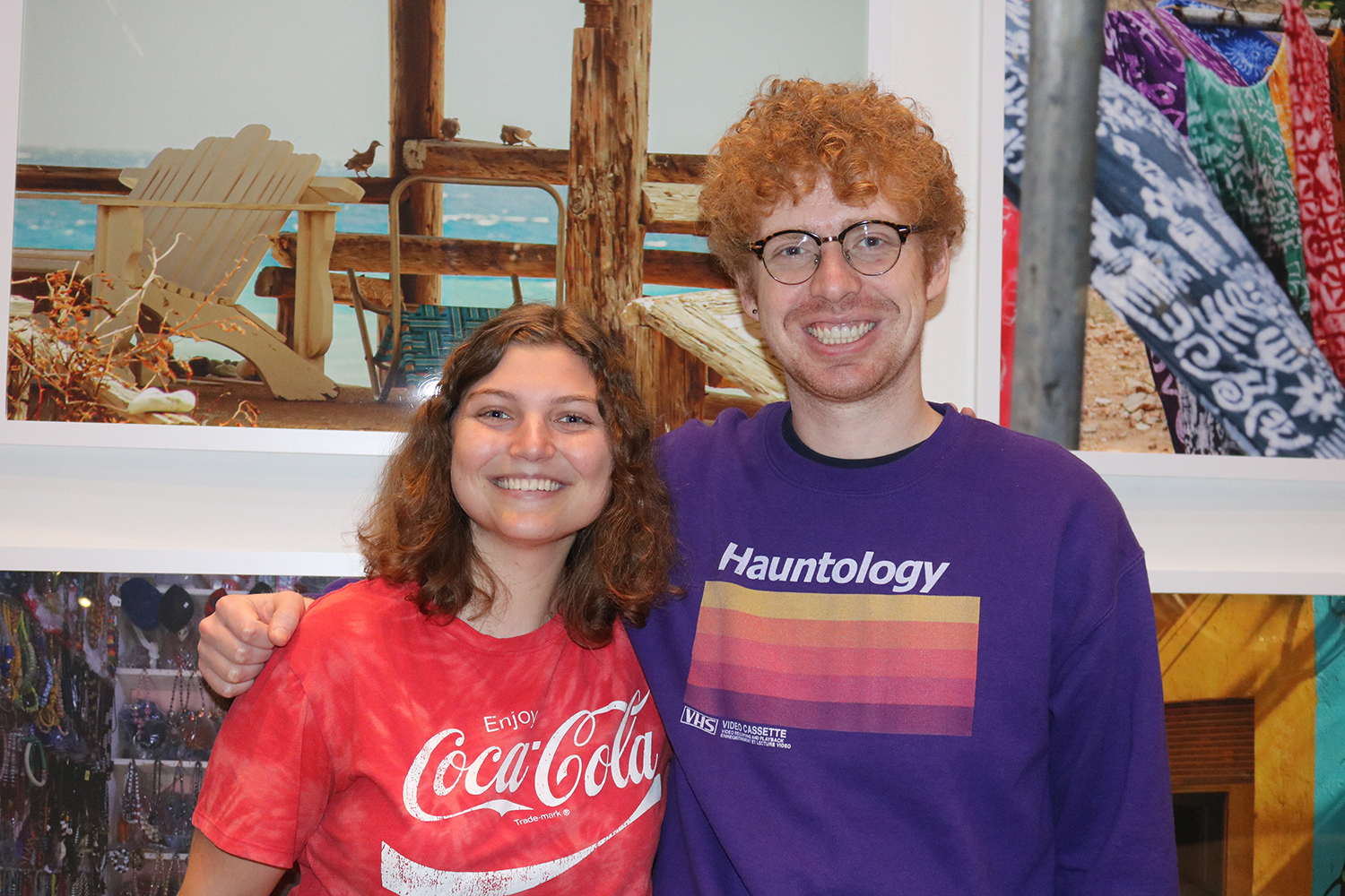 Two individuals pose for a photo in front of colorful artwork in an exhibition, showcasing their casual outfits.