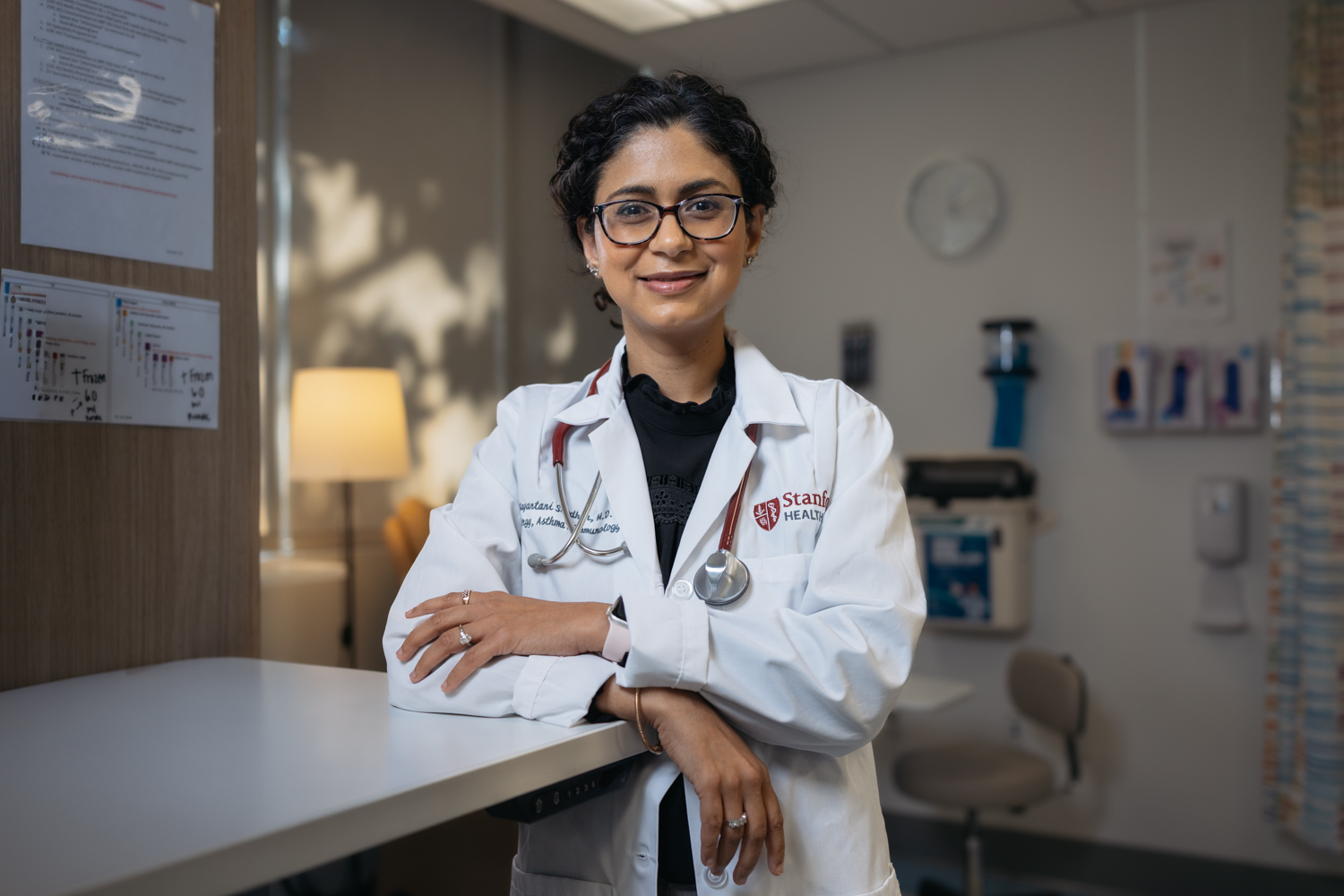 Tina Sindher in a lab coat stands in a clinic, showcasing a patient care environment.