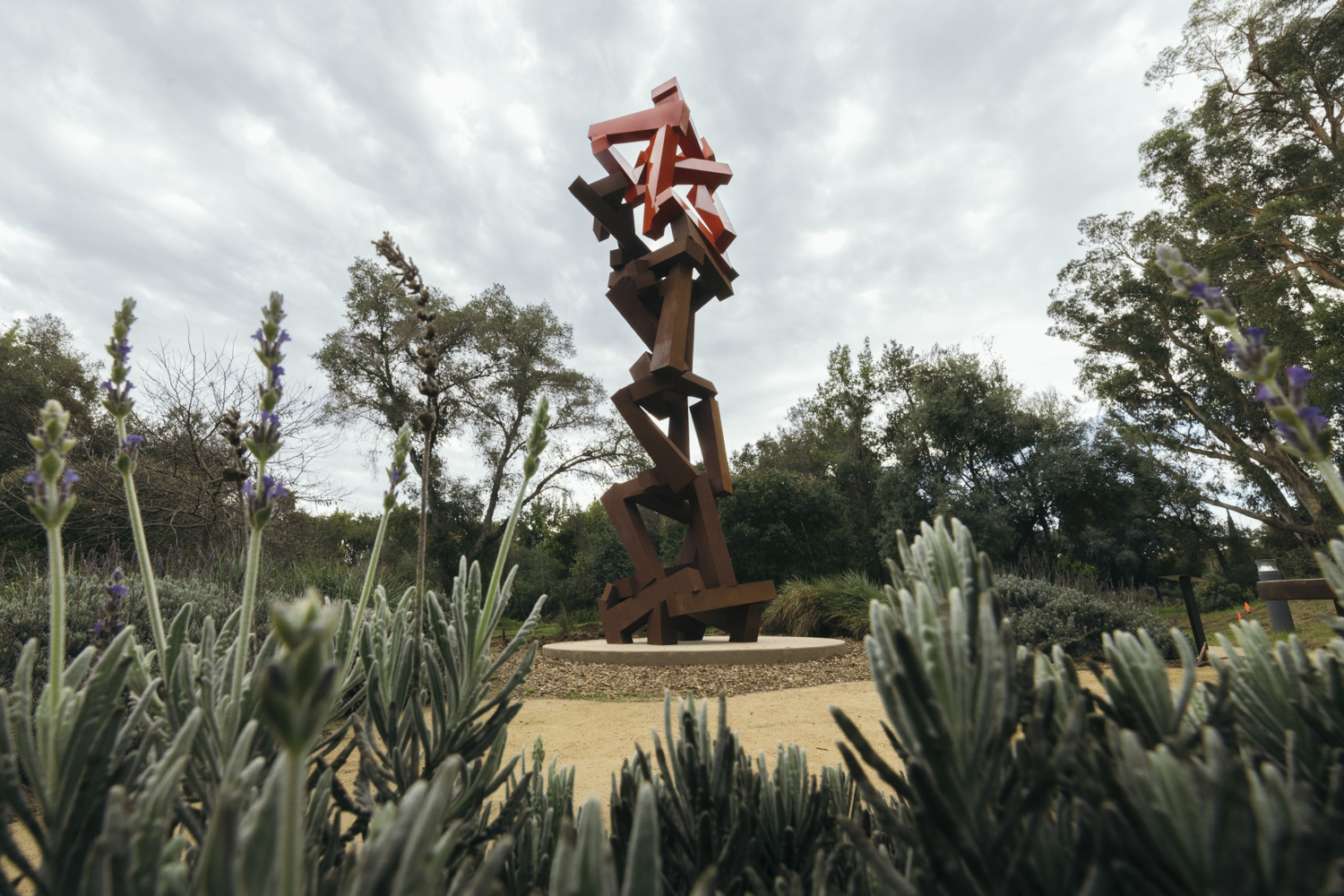 An abstract metal sculpture with red and brown elements is surrounded by lavender plants in a landscaped outdoor area.