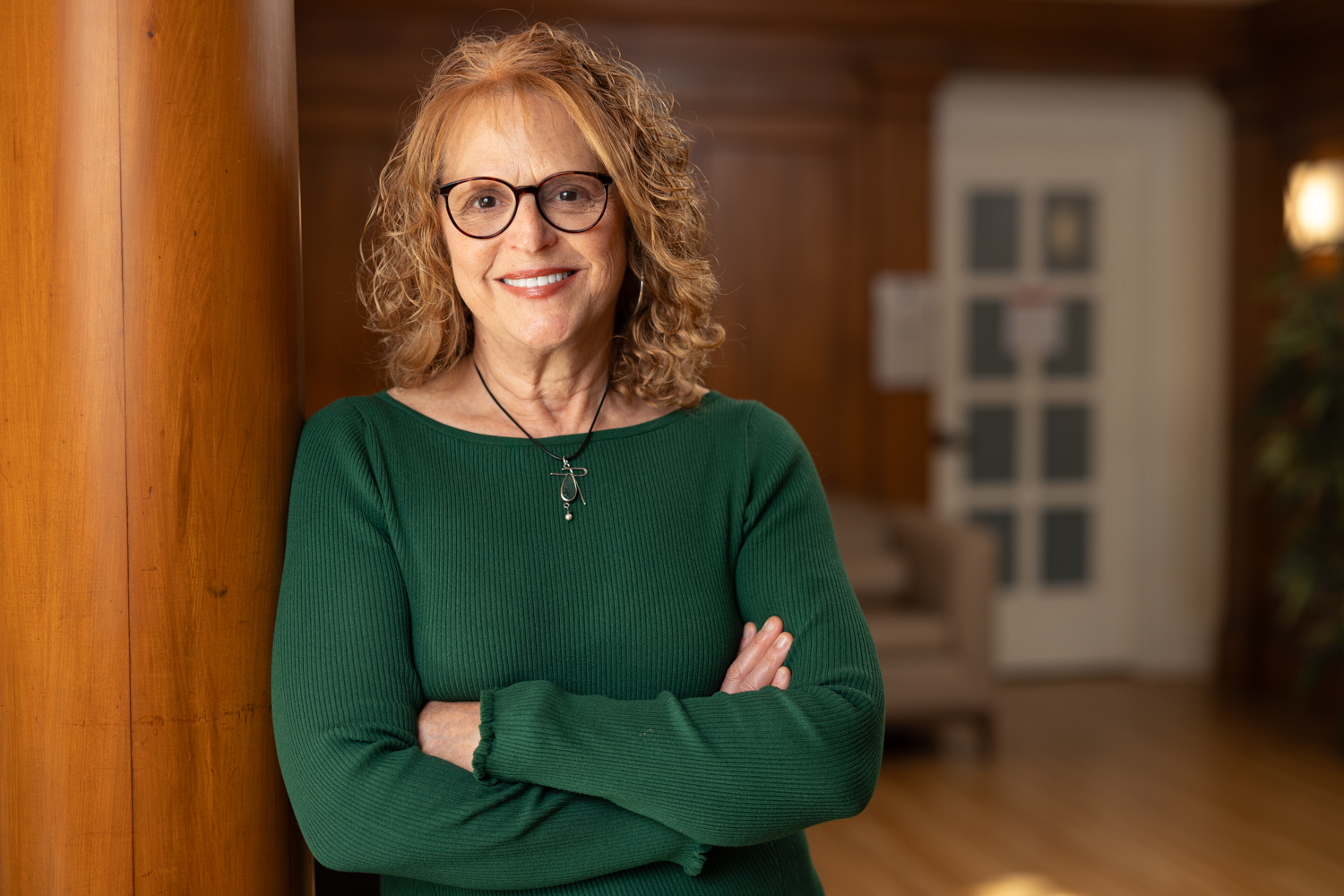 Brenda Berlin stands with her arms crossed, wearing a green sweater, against a wooden backdrop in a well-lit interior.