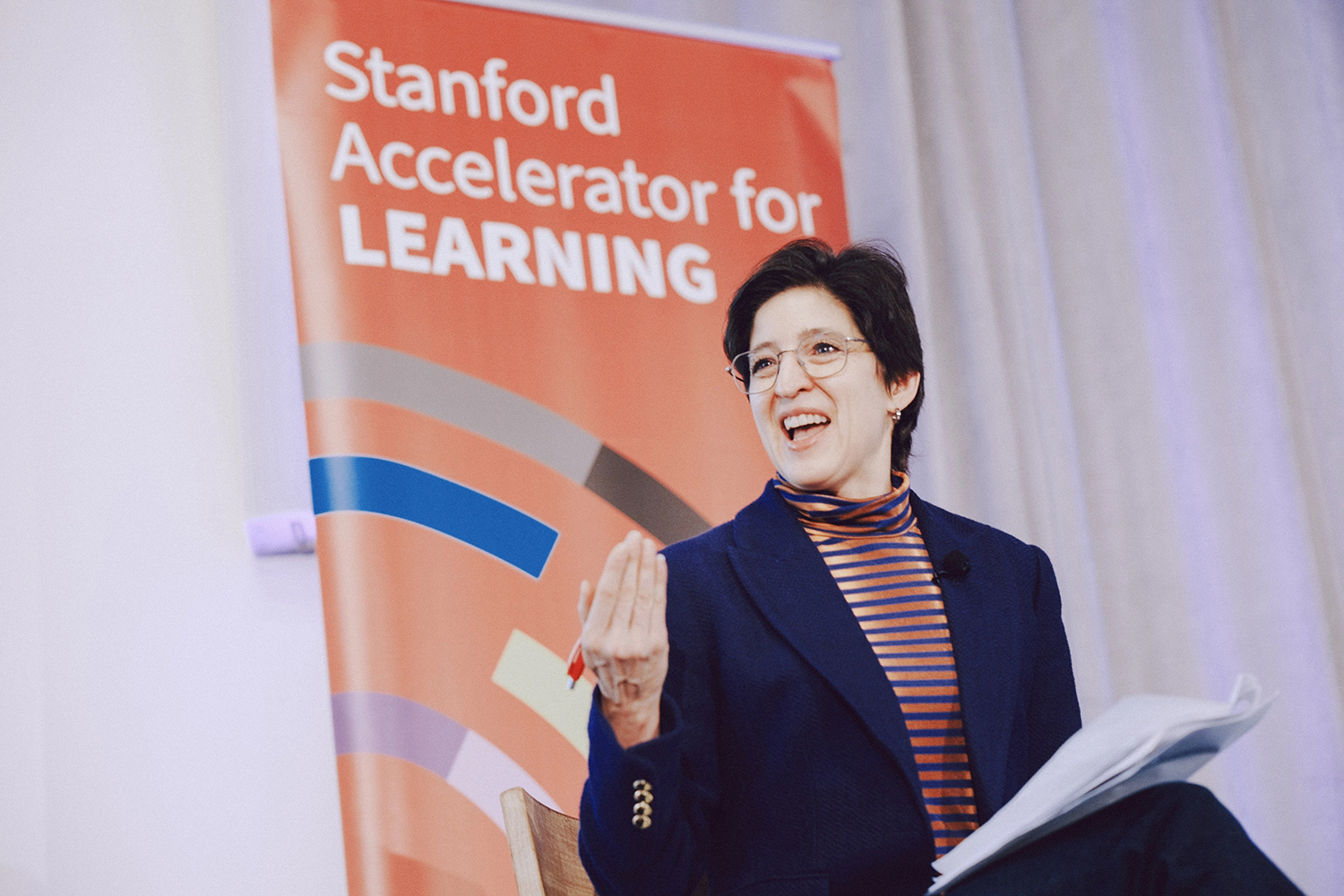 Speaker Susan Athey gestures while sitting next to a banner for the Stanford Accelerator for Learning.