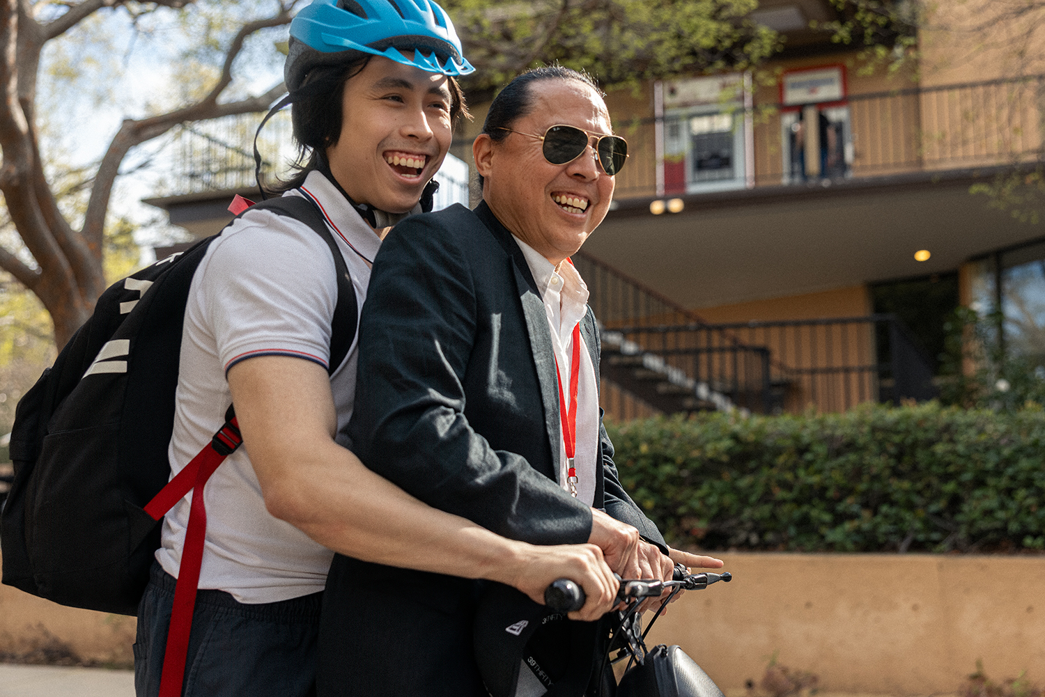 A father and son ride a scooter, one wearing a helmet and casual clothes while the other is dressed in formal attire, in an outdoor setting.
